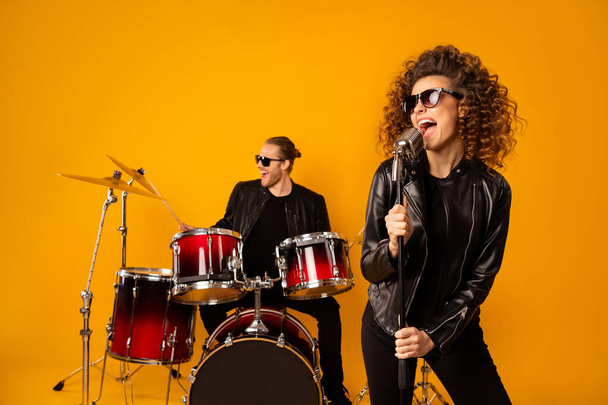Photo of famous rock group guy plays instruments beat drum sticks attractive girl sings mic night club performance show concert hall wear black leather outfit isolated yellow background - Fotó, kép