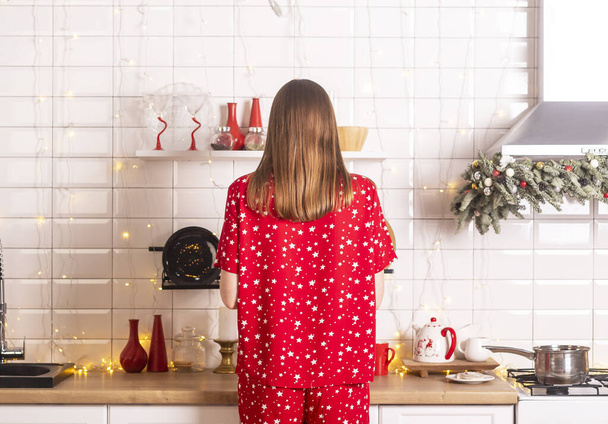 Woman back in pyjamas close up. Cooking festive food at christmas eve in decorated kitchen. Home interior with xmas decoration. Preparing for new year celebration. - Photo, image