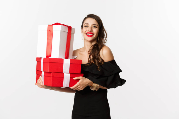 Концепція святкування та різдвяних свят. Модна жінка в чорній елегантній сукні, тримає подарунки та посміхається, стоїть на білому тлі
 - Фото, зображення
