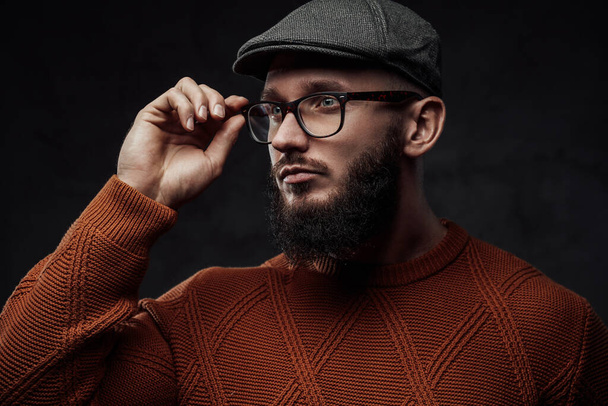 Hipster with beard and cap poses in dark background - Photo, Image