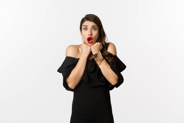 Portrait of glamour woman looking scared and shocked at camera, staring at something with fear, standing in black dress against white background - Photo, Image