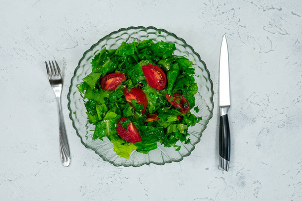 Ensalada vegana verde hecha de una mezcla de hojas verdes y tomates en un tazón de ensalada con un tenedor y un cuchillo en una mesa de piedra blanca. Vista superior sobre gris. - Foto, imagen