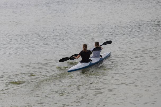 Δύο έφηβες κάνουν καγιάκ στη λίμνη. Πίσω όψη στην ενεργό φάση του εγκεφαλικού. Γραμμένο φόντο - Φωτογραφία, εικόνα