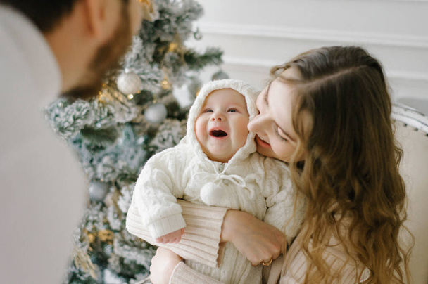 A mother holds a small child in her arms, who smiles at her father. Selective focus on the baby. Happy family celebrates Christmas at the Christmas tree - Photo, Image