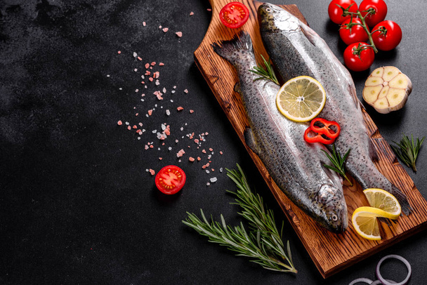 Raw trout lie on a black stone chopping board on a dark concrete table. Nearby are: tomato, rosemary, lemon, pepper, onion, salt - Photo, Image
