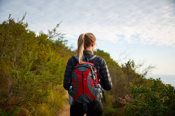 Rear view of young woman on vacation wearing backpack setting off on hike along coastal path next to sea - Photo, Image