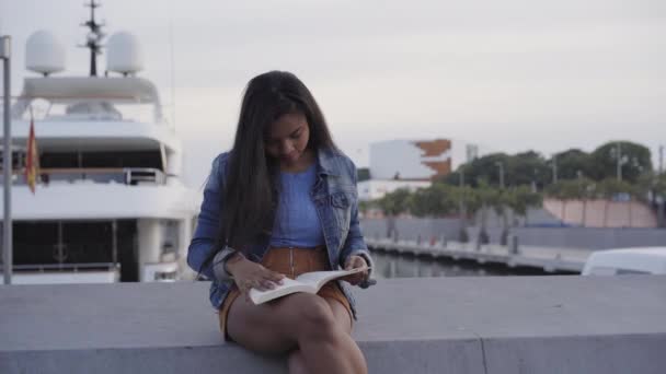 Portrait of an African American black woman reading a book sitting on a bench in the street. Behind her is a harbor with a parked yacht. - Footage, Video