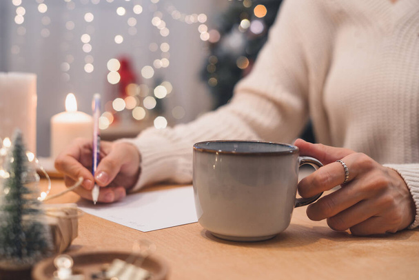 Doelstellingen plannen te doen en verlanglijstje voor nieuwjaar kerst concept schrijven in notebook. Vrouw hand houden pen op notitieblok en koffiekop thuis op wintervakantie xmas. - Foto, afbeelding