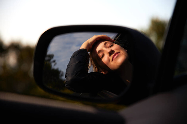 Jovem mulher refletida no espelho dos motoristas com os olhos fechados em férias viagem de carro inclinando-se para fora da janela do carro alugado explorando bela paisagem - Foto, Imagem