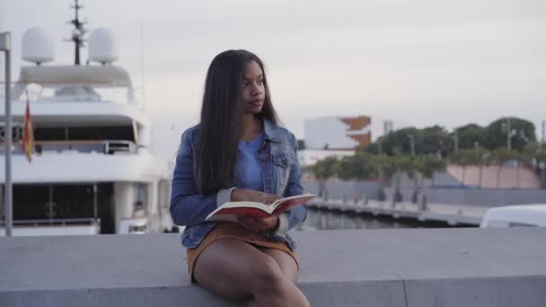 Portret van een zwarte Afro-Amerikaanse vrouw die een boek leest zittend op een bankje op straat. Achter haar is een haven met een geparkeerd jacht. - Video
