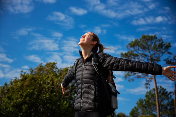 Активная молодая женщина в рюкзаке во время похода по сельской местности раздвигает руки и закрывает глаза, наслаждаясь свободой и пространством. - Фото, изображение
