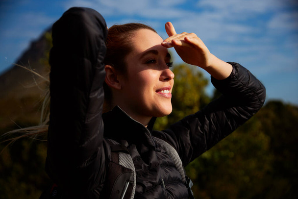 Active young woman wearing backpack on hike in countryside shielding her eyes from the sun - Photo, Image