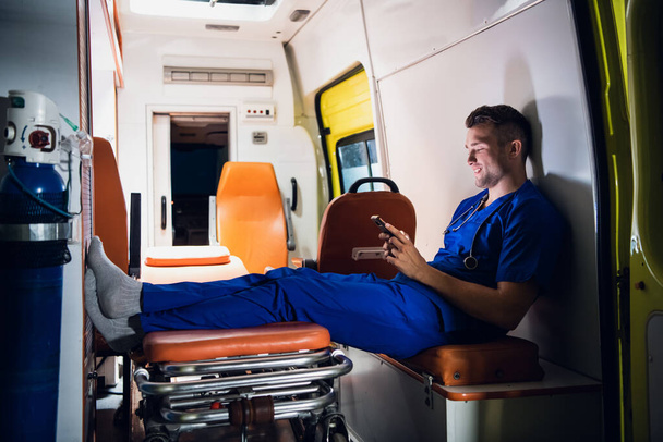 Άντρας τραυματιοφορέας με μπλε στολή ξεκουράζεται και περιηγείται το τηλέφωνό του στο αυτοκίνητο ασθενοφόρο - Φωτογραφία, εικόνα