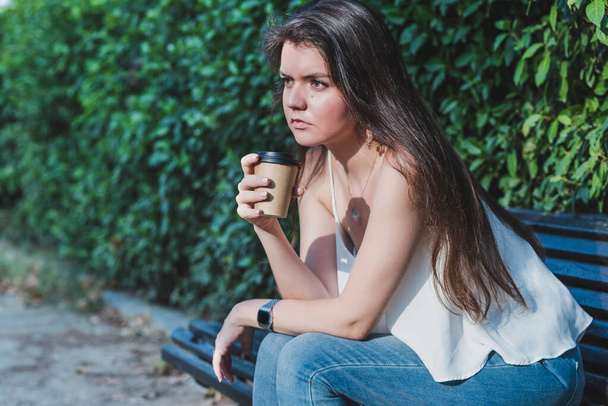 portrait of a young caucasian woman dressing casual with jeans and a white shirt and sitting on a bench of a park with a cup of a paper take away coffee. Selective focus on paper cup - Photo, Image