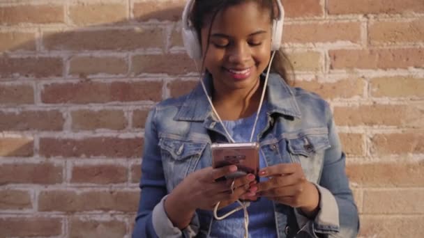 Retrato de una joven afroamericana con auriculares escuchando música, cantando y bailando divertido en una pared de ladrillo. - Imágenes, Vídeo