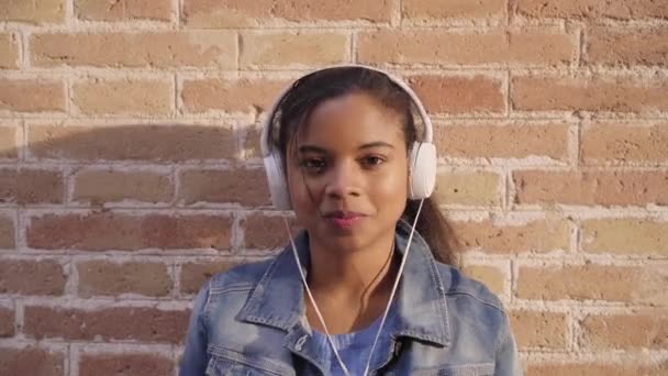 音楽を聴き、歌い、カメラを見てレンガの壁に楽しいダンスをするヘッドフォンを持つ若いアフリカ系アメリカ人女性の肖像画. - 映像、動画