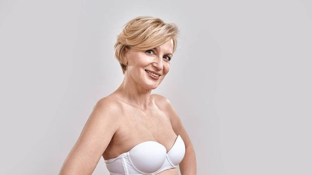 Portrait of beautiful middle aged woman in white bra smiling at camera, posing isolated against grey background. Beauty concept - Фото, изображение