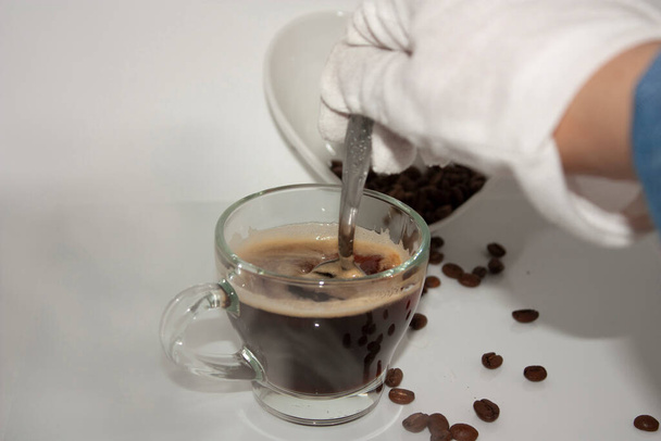 Tasse à vapeur avec café et mousse se déplaçant par cuillère en remuant à l'aide d'une main blanche gantée, à côté d'un boulon en céramique blanche en forme de coeur - Photo, image