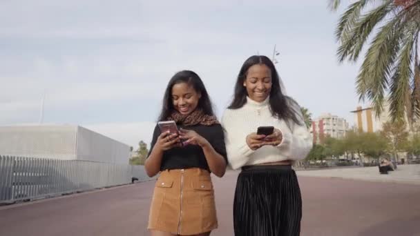 Twee zwarte meisjes die in een stad lopen terwijl ze een mobiele telefoon gebruiken en lachen. Mensen en technologie concept met smartphone. - Video