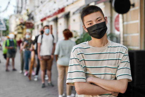 Portrait of young asian guy wearing mask waiting, respecting social distancing to collect his takeaway order from the pickup point during lockdown - 写真・画像