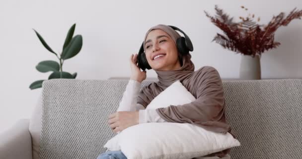 Jonge vrolijke moslim dame genieten van favoriete muziek, zitten op de bank met kussen dragen hoofdtelefoon en ontspannen - Video