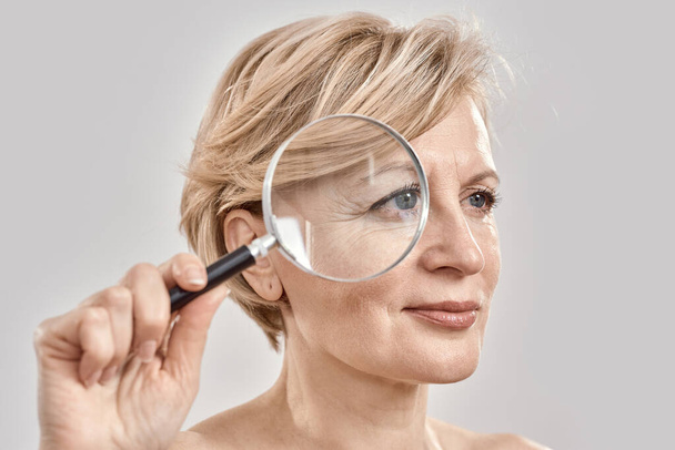 Close up portrait of attractive middle aged woman looking aside, holding a magnifying glass and showing her wrinkles, posing isolated over grey background - Zdjęcie, obraz