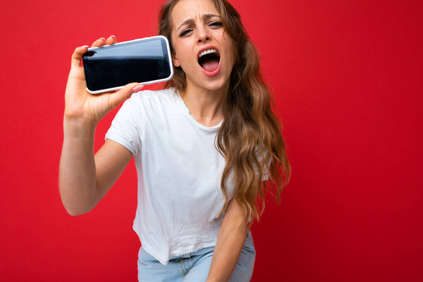 Afbeelding van een geschokte jonge mooie vrouw poseren geïsoleerd over rode muur achtergrond spelen games door mobiele telefoon. - Foto, afbeelding