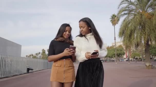 Twee zwarte meisjes die in een stad lopen terwijl ze een mobiele telefoon gebruiken en lachen. Mensen en technologie concept met smartphone. - Video