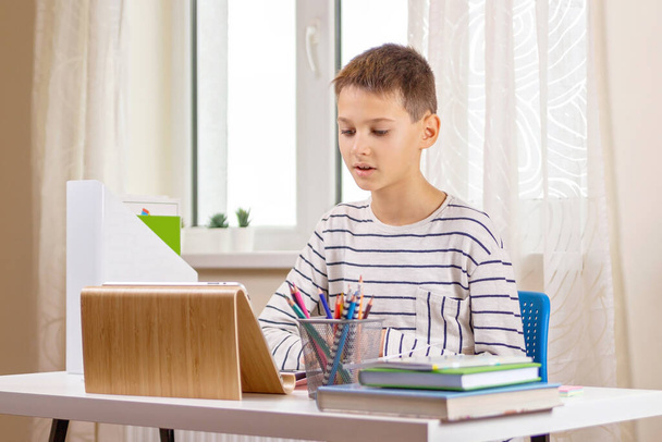 Kid tabletin tietokone istuu pöydässä kirjoja ja ottaa videopuhelu, virtuaalinen online leasson kotona - Valokuva, kuva