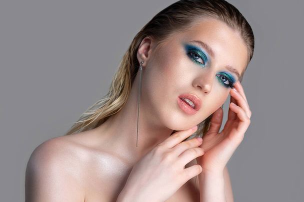 Closeup studio portrait of an attractive blonde woman with wet hair and deep blue smokey eyes makeup. Model posing with naked shoulders over a grey background. Empty space - Фото, изображение