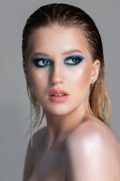 Closeup studio portrait of a seductive blonde woman with wet hair and deep blue smokey eyes makeup. Model posing with naked shoulders over a grey background - Photo, image