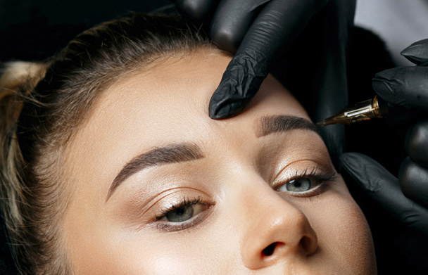 Cosmetician in gloves making brow permanent makeup to a stunning blonde woman in a beauty salon. Top view. Closeup sho - Photo, Image