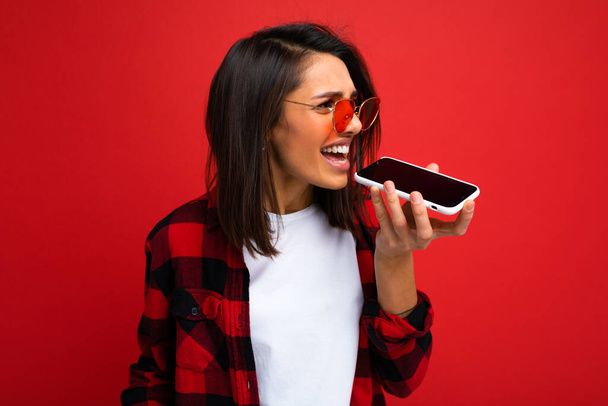 brunette on the background of a red wall speaks on the speakerphone on the phone, angry  - Photo, Image