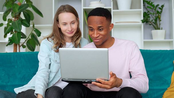 Cheerful interracial couple web surfing on laptop together, sitting on sofa at home - Foto, Imagem