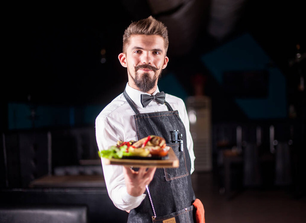 Professional friendly waiter helpfully serves plate with prepared dish on a black background. - Photo, Image