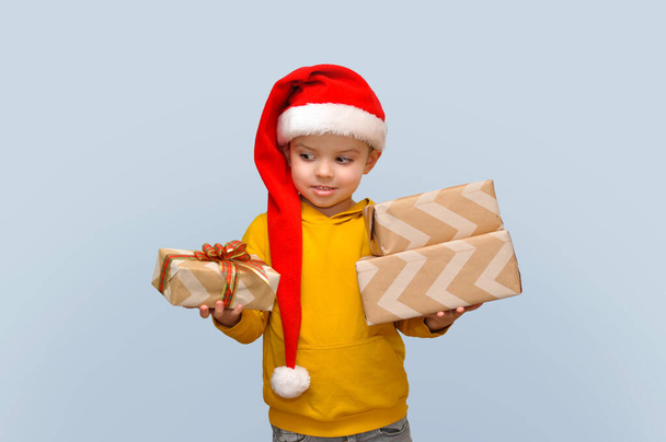 A cute boy in a santa hat holds a lot of craft boxes with gifts. - Fotografie, Obrázek