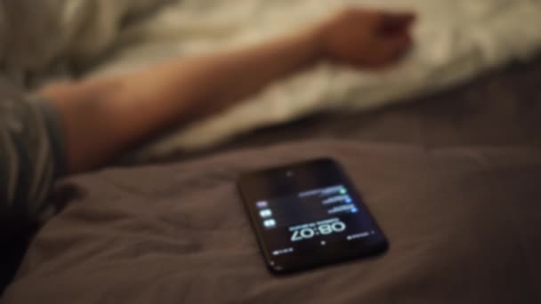 早朝に携帯の目覚まし時計で目を覚ます若い男。コンセプト。スマートフォンの目覚まし時計の信号を聞いた後に目を覚ます眠そうな男は、携帯電話を鳴らすために到達し、それを使用して. - 映像、動画