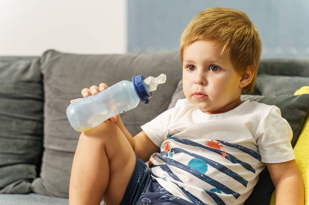 Small caucasian boy holding baby bottle while sitting on the bed at home looking to the side alone - Photo, Image