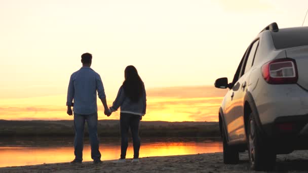Feliz pareja de viajeros se paran junto al coche y admiran la hermosa puesta de sol sobre el río en la playa. turistas al lado de coche, admirando la salida del sol, lago. Viajeros libres, turistas. viajes familiares en coche. - Imágenes, Vídeo