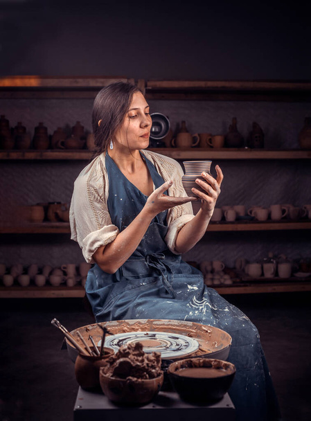Κομψή τεχνίτης γυναίκα κάθεται στον πάγκο με κεραμικό τροχό και την κατασκευή πήλινη κατσαρόλα. Χειροποίητα προϊόντα. - Φωτογραφία, εικόνα