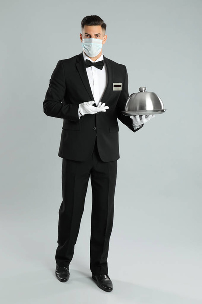 Camarero con mascarilla médica sosteniendo bandeja sobre fondo gris claro - Foto, imagen