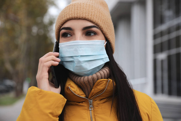Young woman in medical face mask talking on phone while walking outdoors. Personal protection during COVID-19 pandemic - Valokuva, kuva
