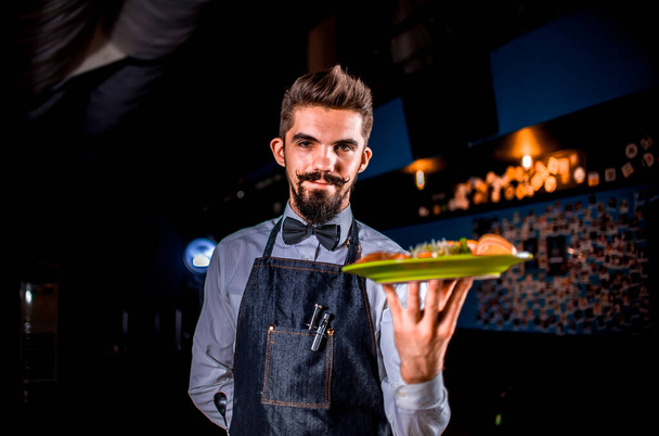 Profesjonalny przyjazny kelner pomocnie serwuje dania gotowane na czarnym tle. - Zdjęcie, obraz