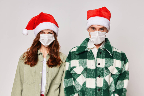 Ένα χαριτωμένο νεαρό ζευγάρι με ιατρικές μάσκες στέκεται δίπλα σε ένα κοντινό καπέλο του Άη Βασίλη. - Φωτογραφία, εικόνα