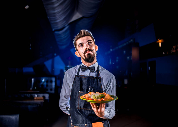 Νεαρός σερβιτόρος κρατά σαλάτα σε μαύρο φόντο. - Φωτογραφία, εικόνα