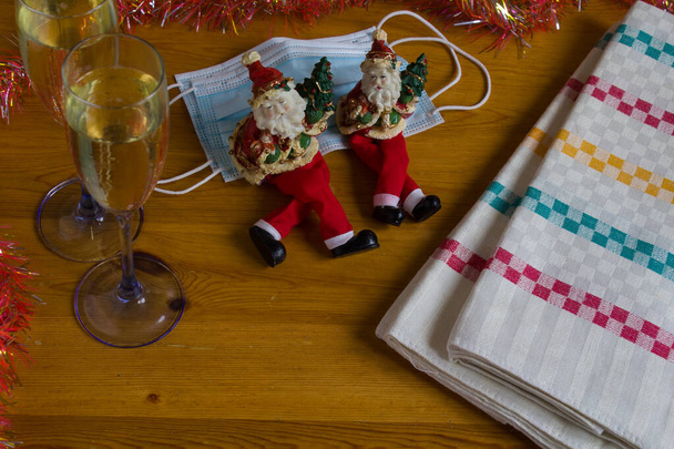 Concept van Kerstmis en Nieuwjaar met coronavirus.Champagne glazen op een houten tafel met maskers voor de gezelligheid19. Er zitten twee kerstmanfiguren comfortabel. Met typische decoratie. - Foto, afbeelding