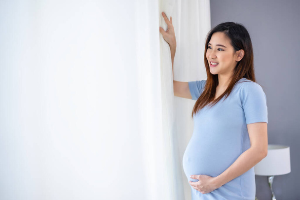 happy pregnant woman looking view at window and stroking her belly in the bedroo - Photo, image