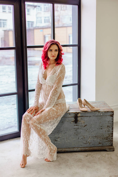 Attractive woman with pink hair in white light dress sitting on the old wooden box near large window. Lady in gentle lace peignoir sitting with bare feet in a room with concrete floor - Photo, Image
