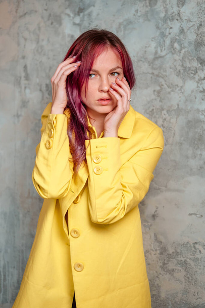 Beautiful woman with pink hair dressed in yellow raincoat touching her face and posing indoor near the concrete wall. - Foto, imagen