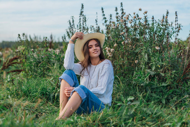 Fiatal gyönyörű barna nő öltözött fehér pulóver, farmer és cowboy szalma kalap ül a zöld fű, mosolyog és nevet napnyugtakor - Fotó, kép